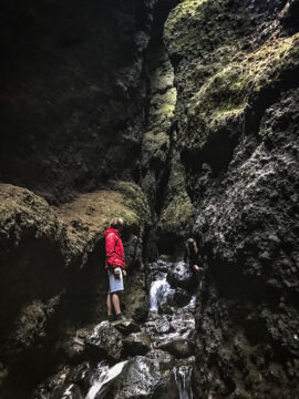 A hiker in a red jacket standing on rocks by the stream inside the dark Rauðfeldsgjá crevice