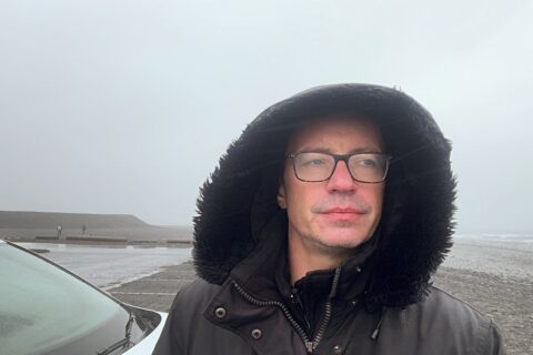 Author standing on Diamond Beach Iceland in horizontal rain, soaked jacket, grey skies and waterlogged parking lot behind