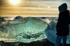 Person examining a naturally fish-shaped piece of glacial ice on Diamond Beach Iceland, sunlight shining through the ice with sparkling reflections, ocean in the background