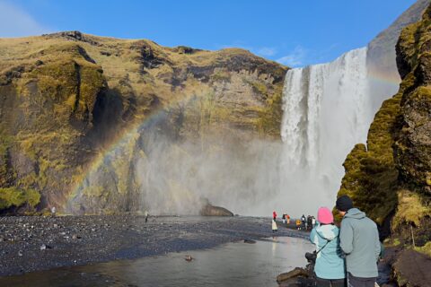 Couple looking at Skógafoss waterfall on a sunny autumn day in Iceland, with a rainbow formed in the mist and clear blue sky above